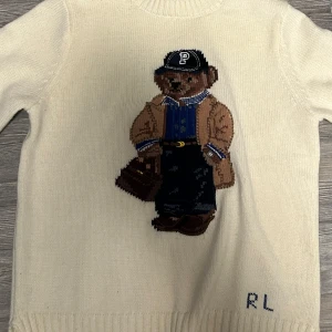  Stickad Polo bear - Säljer en vit stickad tröja från Polo Ralph Lauren med ett coolt björnmotiv på framsidan. Tröjan har en klassisk rund hals och långa ärmar. Perfekt för en avslappnad och stilren look.