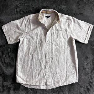 Burberry skjorta  - Säljer en stilren vit kortärmad skjorta från Burberry. Skjortan har en klassisk krage med det ikoniska Burberry-mönstret på insidan. Den är försedd med knappar och en broderad logotyp på bröstfickan. Perfekt för en elegant och avslappnad look. Skick 8/10, storlek M men passar även L. Pris går att diskutera✅