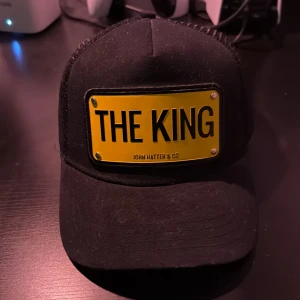 Svart keps från John Hatter & Co - Snygg svart keps med en gul skylt framtill där det står 'THE KING'. Märket John Hatter & Co är designat i Sverige. Perfekt för att ge en cool touch till din outfit.