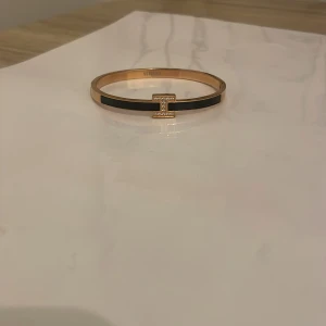 Elegant armband med H-design - Snyggt armband i guld med en svart detalj och en H formad design prydd med små stenar. Perfekt för att ge en stilren touch till din outfit.
