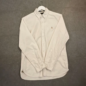 Vit skjorta från Ralph Lauren - Snygg vit skjorta från Ralph Lauren med broderad logotyp på bröstet. Skjortan har en slim fit passform och är långärmad med knappar hela vägen ner. Perfekt för en stilren look.