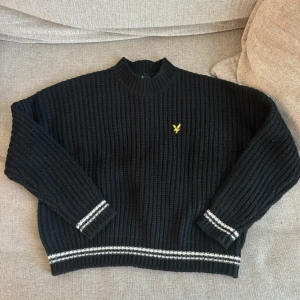 Svart stickad tröja från Lyle & Scott - Säljer en svart stickad tröja från Lyle & Scott med ribbade detaljer och ett gult logomärke på bröstet. Tröjan har långa ärmar och vita ränder vid ärmslut och nederkant. Perfekt för kyliga dagar! Lite nopprig som man kan se på sista bilden 