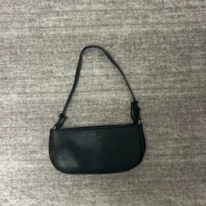 Svart axelväska från NA-KD - Snygg svart axelväska från NA-KD i skinnmaterial. Väskan har en stilren design med en praktisk dragkedja upptill. Perfekt för att bära dina nödvändigheter med stil.