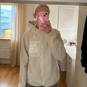 Beige fleecejacka med dragkedja - Mysig beige fleecejacka med dragkedja och huva. Jackan har en praktisk bröstficka med ett tryckt märke. Perfekt för kyliga dagar och ger en avslappnad look.