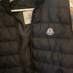 Svart dunväst från Moncler - Snygg svart dunväst från Moncler med quiltad design och dragkedja. Perfekt för kyligare dagar och ger en stilren look. Moncler-logga broderad på bröstet. Storlek L. Är 188 lång och väger 80 kg och den passar mig bra