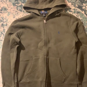 Grön hoodie från Ralph Lauren - Säljer en stilren grön hoodie från Ralph Lauren med dragkedja och huva. Tröjan har långa ärmar och en liten broderad logga på bröstet. Perfekt för en avslappnad look.