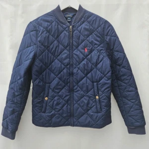 Blå quiltad jacka från Ralph Lauren - Snygg blå quiltad jacka från Ralph Lauren med dragkedja och broderad logga på bröstet. Jackan har långa ärmar och praktiska fickor med knappar. Perfekt för kyligare dagar. Storlek L i barn så ca S