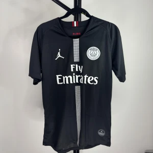 Svart PSG fotbollströja - Snygg svart PSG fotbollströja med Fly Emirates-logga och Jordan-märke. Tröjan har en stilren design med korta ärmar och en diskret randig detalj i mitten. Perfekt för fotbollsfans som vill ha en sportig look.