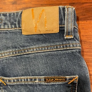 Blå jeans från Nudie Jeans - Snygga blå jeans från Nudie Jeans med klassisk design och orange sömmar. Perfekta för en avslappnad stil. Märkeslogga på bakfickan och läderpatch i midjan.