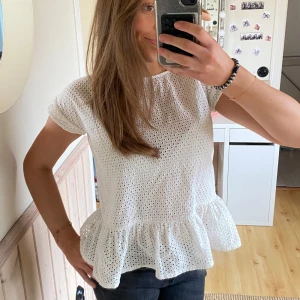 Vit blus med volangdetalj - Säljer en söt vit blus med korta ärmar och volangdetalj i nederkant. Blusen har ett hålmönster som ger en luftig känsla. Perfekt för en avslappnad och stilren look.