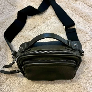 Väska - Grå väska med axelband. Har två fack framme och ett litet bak. Använd få gånger. Väldigt snygg!