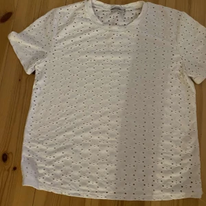 Vit t-shirt från Zara - Säljer en snygg vit t-shirt från Zara. Den är i storlek m och har ett coolt hålmönster över hela tyget. Perfekt för sommaren eller som en del av en lager-på-lager outfit. T-shirten är i bra skick och väldigt bekväm att ha på sig. Passar både till vardags och fest!