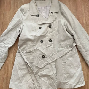 Beige trenchcoat med bälte - Säljer en snygg beige trenchcoat i storlek M. Jackan har  dubbelknäppning och ett bälte i midjan för en figursydd passform. En av knapparna saknas (bild 1)  påverkar inte användningen och märks knappt då man fortfarande kan stänga kappan. man fortfarande Perfekt för hösten🥰