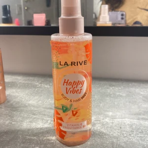 Happy Vibes Body & Hair Mist från La Rive - Säljer en fräsch och upplyftande body & hair mist från La Rive med namnet Happy Vibes. Flaskan är genomskinlig med en orange etikett och färgglada detaljer. Den innehåller vitamin E och provitamin B5, vilket gör den perfekt för att återfukta både hud och hår. Flaskan rymmer 200ml (6.7 fl oz) och har en praktisk spraypump för enkel applicering. Perfekt för att ge en fräsch doft och känsla under dagen!