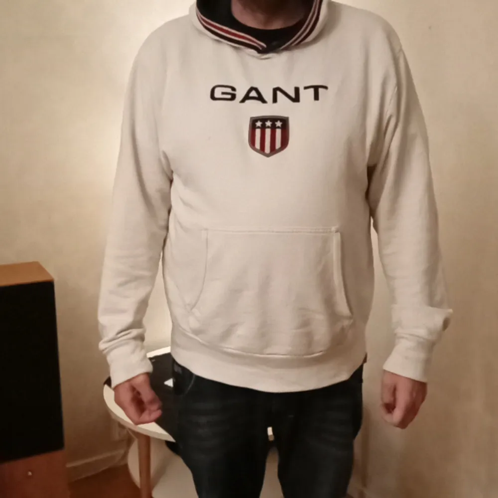 Säljer en snygg vit hoodie från GANT. Den har en broderad logga på bröstet och en känguruficka framtill. Hoodien har långa ärmar och en ribbad mudd vid ärmslut och nederkant. Perfekt för höst och vinter!. Tröjor & Koftor.