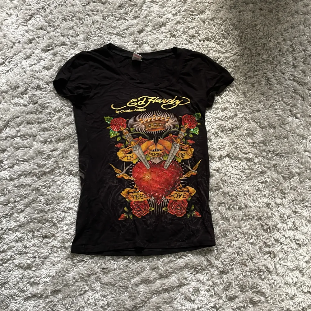 Säljer en svart Ed Hardy t-shirt i storlek S. Den har ett coolt tryck med ett stort hjärta och rosor på framsidan, och tigrar och en dödskalle på baksidan. T-shirten är kortärmad och gjord av bomull. Perfekt för en casual look eller för att sticka ut på festen! 🖤. T-shirts.