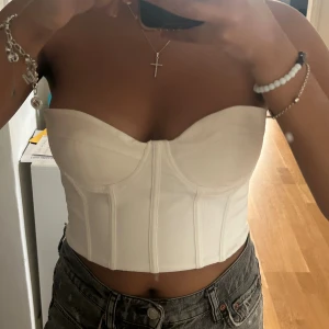 Vit korsett-topp - Säljer en snygg vit korsett-topp i storlek S. Den är axelbandslös och har en tight passform som framhäver figuren. Perfekt för fest eller en utekväll! Toppen är i nyskick och har en stilren design med sömmar som ger en extra touch. Tröjan har en dragkedja så det blir enkelt att sätta på och av men dragkedjan syns inte utåt.