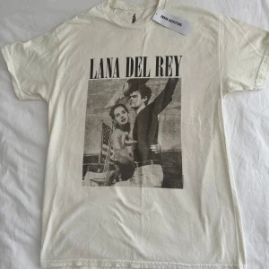 Vit t-shirt med Lana Del Rey tryck från Urban Outfitters - Säljer en vit t-shirt från Urban Outfitters med ett stort svartvitt tryck av Lana Del Rey på framsidan. T-shirten är i oversized fit och är gjord av bomull. Perfekt för alla Lana Del Rey-fans som vill ha en cool och bekväm t-shirt att bära till vardags.