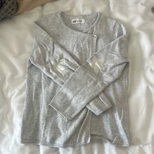 Grå stickad kofta från H&M - Säljer en mysig grå stickad kofta från H&M. Den har långa ärmar och en fin silverstjärna som detalj på armbågarna. Koftan har en knapp framtill för att hålla den stängd. Perfekt för höst och vinter!