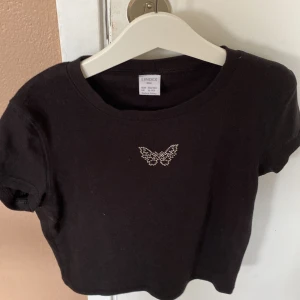 Svart croppad topp med fjärilsdetalj från Lindex - Säljer en svart croppad topp från Lindex med en söt fjärilsdetalj i pärlor på bröstet. Toppen är kortärmad och gjord i bomull, perfekt för en avslappnad och trendig look. strl 154-164 köparen betalar för frakt!