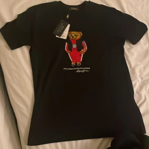 Svart t-shirt från Polo Ralph Lauren - Säljer en helt ny svart t-shirt från Polo Ralph Lauren med en cool broderad design på framsidan. T-shirten är i nyskick och har korta ärmar. Perfekt för en avslappnad och stilren look!