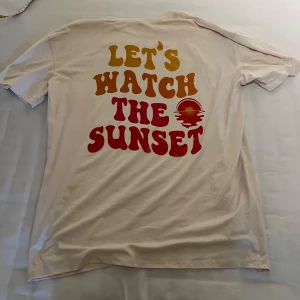 Vit t-shirt med texten 'Let's Watch The Sunset' - Säljer en vit t-shirt i storlek L med en cool retroinspirerad text 'Let's Watch The Sunset' i gult och rött. T-shirten är kortärmad och perfekt för sommaren. Den är i bra skick och passar både till vardags och fest. Materialet känns som bomull, vilket gör den skön att ha på sig. Perfekt för dig som vill ha en avslappnad och stilren look!
