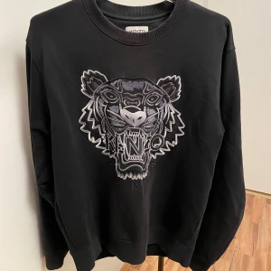 Kenzo tröja - Snygg svart sweatshirt från Kenzo med ett stort, detaljerat tigerbroderi på framsidan. Tröjan har rund hals och långa ärmar, perfekt för en stilren look. Passar bra till både jeans och chinos. 