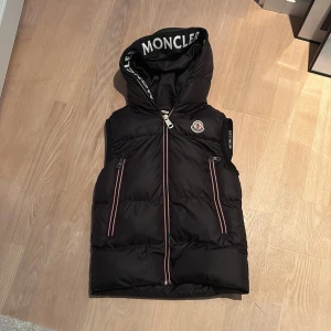 Svart dunväst från Moncler för barn - Snygg svart dunväst från Moncler med huva och dragkedja. Västen har två fickor med dragkedjor och Moncler-logga på bröstet. Perfekt för kyligare dagar.