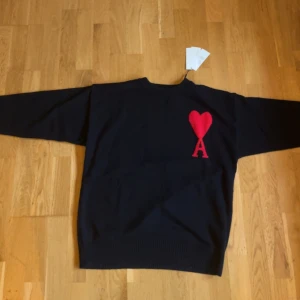 Svart tröja från Ami Paris - Säljer en snygg svart tröja från Ami Paris med ett rött hjärta och bokstaven A på bröstet. Tröjan har långa ärmar och en klassisk passform. Perfekt för en stilren look. Kan sänka pris vid snabb affär             TAR EMOT BYTEN