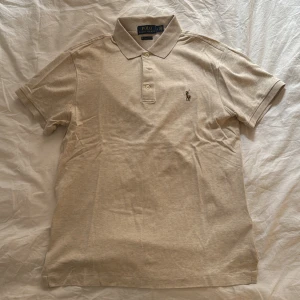 Pikétröja från Ralph Lauren - Beige pikétröja från Ralph Lauren i slim fit. Tröjan har korta ärmar och klassisk krage med knappar. Den ikoniska broderade loggan syns på bröstet. Använd 1-2 gånger. Nypris ca 1000kr