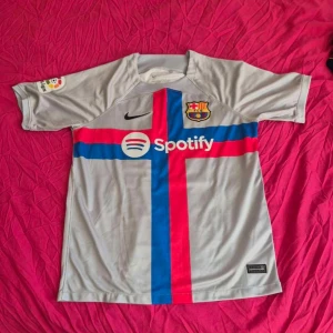Barcelona fotbollströja  - Säljer en grå fotbollströja från Nike med korta ärmar. Tröjan har ett stort kors i blått och rött på framsidan och ett klubbmärke på bröstet. På baksidan finns namnet och numret 'F. DE JONG 21'. Perfekt för fotbollsfans!
