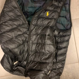 Svart dunväst från Polo Ralph Lauren - Snygg svart dunväst från Polo Ralph Lauren med dragkedja och broderad logga i gult på bröstet. Perfekt för kyligare dagar när du vill hålla stilen. Västen har en quiltad design och praktiska fickor med dragkedja.