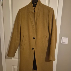 Beige kappa från Zara - Snygg beige kappa från Zara med stilrena knappar framtill. Den har en klassisk design med långa ärmar och en rak passform. Perfekt för att hålla stilen under kyligare dagar.