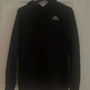 Svart hoodie från Kappa - Säljer en stilren svart hoodie från Kappa med deras ikoniska logga på bröstet. Tröjan har en klassisk design med huva och långa ärmar. Perfekt för en avslappnad look.