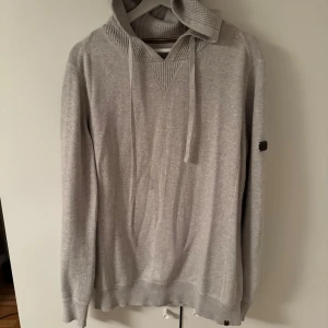 Grå kashmirblandning hoodie  med ribbade detaljer - Säljer en stilren grå hoodie med ribbade muddar och nederkant. Tröjan har en huva med snörning och en diskret logga på ärmen. Perfekt för en avslappnad look.