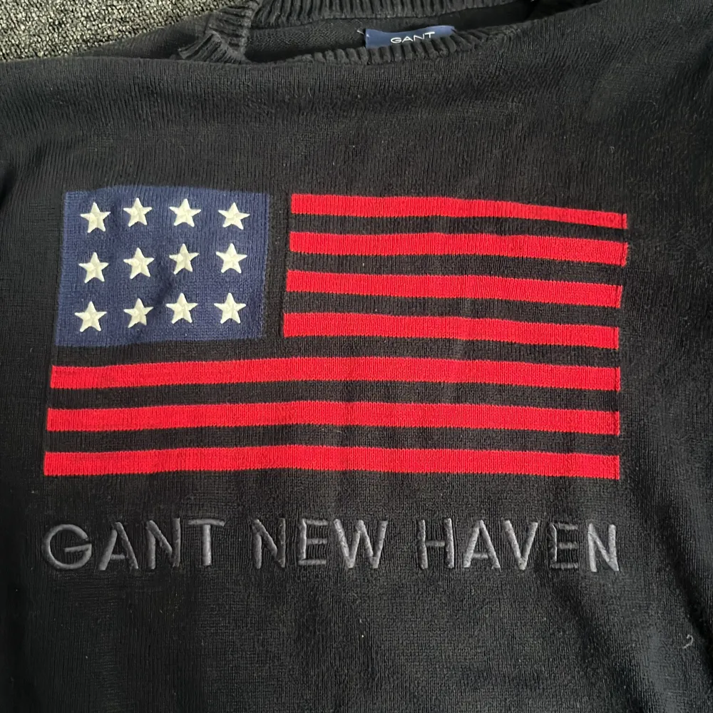 Snygg svart tröja från GANT med ett stort flaggmotiv i rött, vitt och blått på ryggen. Texten 'GANT NEW HAVEN' är tryckt under flaggan. Perfekt för en stilren och avslappnad look.. Neuletakit & Villapaidat.