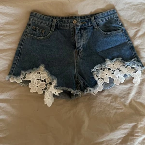 Jeansshorts med spetsdetaljer - Snygga jeansshorts med vackra spetsdetaljer vid benen. Perfekta för sommardagar när du vill ha en avslappnad men stilren look. De har klassiska fem fickor och en knappgylf.