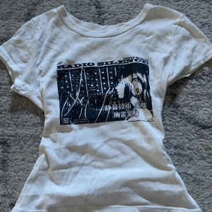 Vit t-shirt med tryck från John Galt - Säljer en vit t-shirt från Brandy Melville med ett coolt svart tryck på framsidan. T-shirten är kortärmad och tillverkad i 100% bomull, vilket gör den bekväm att bära. 