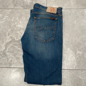  Nudie Jeans tight long John - Snygga blå jeans från Nudie Jeans med klassisk femficksdesign och orange sömmar. De har en knappgylf och är tillverkade i slitstarkt denim. Perfekta för en avslappnad stil. Modellen är tight long John 