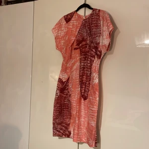 Mönstrad klänning från Acne - Snygg klänning från Acne i en mix av rött och rosa med ett unikt mönster. Klänningen har korta ärmar och en dragkedja bak. Storlek 38, men känns mer som en 36a.