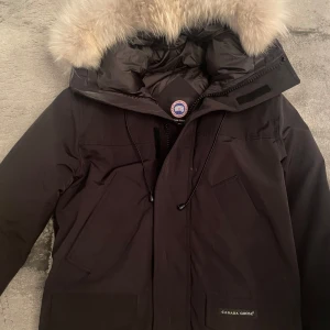 Svart dunjacka från Canada Goose - Säljer en svart dunjacka från Canada Goose med pälskantad huva. Jackan har dragkedja och flera praktiska fickor. Perfekt för kalla vinterdagar.