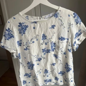 Vit blommig topp från Mango - Säljer en söt vit topp från Mango med blått blommönster. Toppen har korta ärmar och en liten bröstficka. Den har en knappdetalj i nacken för enkel påklädning. Perfekt för en somrig look!