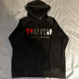 Svart hoodie från Trapstar - Säljer en svart hoodie från Trapstar med texten 'Trapstar It's a Secret' i rött, grått och vitt på framsidan. Hoodien har en klassisk design med dragsko i huvan och en stor magficka. Perfekt för en avslappnad stil.