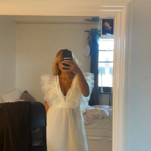 Vit klänning med volangdetaljer (studentklänning) - Säljer en elegant vit klänning med volangdetaljer på axlarna och en v-ringad design. Perfekt för sommarens festligheter. Klänningen har en luftig passform och är tillverkad i ett lätt material.