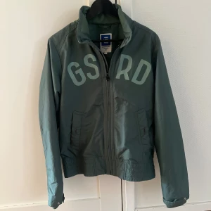 Grön jacka från G-Star RAW - Snygg grön jacka från G-Star RAW med stor text 'GSRD' på framsidan. Jackan har dragkedja och ribbade detaljer vid ärmslut och nederkant. Perfekt för en stilren look.