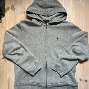 Grå hoodie från Polo Ralph Lauren - Barn storlek,Säljer en klassisk grå hoodie från Polo Ralph Lauren med dragkedja och broderad logga på bröstet. Tröjan har en bekväm passform och är perfekt för kyligare dagar. Den har långa ärmar och en praktisk huva.