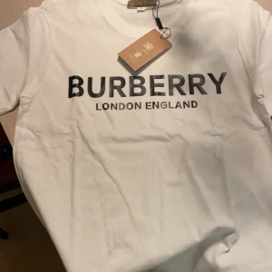 Vit t-shirt från Burberry - Snygg vit t-shirt från Burberry med svart text 'BURBERRY LONDON ENGLAND' på framsidan. Perfekt för en stilren och klassisk look. T-shirten har en rund hals och korta ärmar.