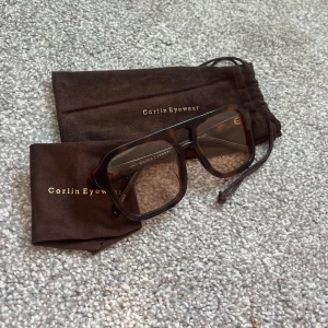 Bruna glasögon från Corlin Eyewear - Säljer ett par snygga glasögon från Corlin Eyewear. De har en fyrkantig form och kommer i en stilren brun färg. Perfekt för att ge din outfit en extra touch! Glasögonen kommer med en matchande brun påse för förvaring.
