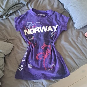 Lila t-shirt med tryck från Way Nor - Säljer en cool lila t-shirt från Way Nor i storlek XS. Den har ett stort 'NORWAY'-tryck i vitt på framsidan och dekorativa mönster i rött och vitt. Perfekt för en avslappnad stil eller som souvenir. T-shirten är kortärmad och gjord i ett mjukt material.