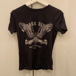 Svart t-shirt från Gina Tricot - Säljer en cool svart t-shirt från Gina Tricot i storlek XS. Den har ett tryck med texten 'Arizona Desert' och en bild av en örn med utsträckta vingar. Perfekt för en avslappnad stil!